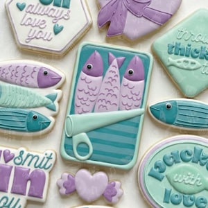May include: An assortment of decorated cookies in various shapes and colors. The cookies include fish, a sardine can, hearts, and cookies with text like "always love you" and "packed with love". The color palette includes shades of blue, purple, and white.