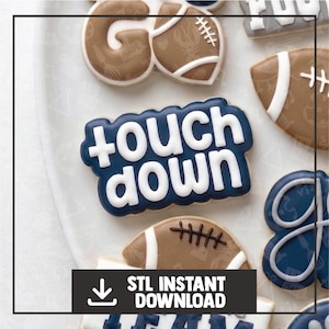 May include: A collection of decorated sugar cookies featuring a dark blue plaque with the white text "touch down" in a bold font, alongside tan football-shaped cookies with white laces and a partial cookie with a blue and white design.