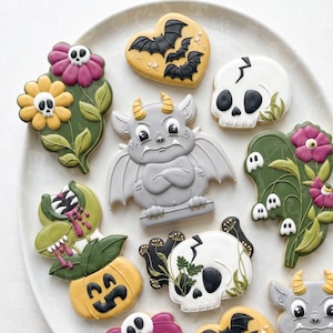 May include: A collection of decorated sugar cookies on a plate, featuring a gray gargoyle, white skulls with black details, a yellow heart with black bats, a pumpkin with a plant, and flowers with skull centers, all with intricate icing designs.