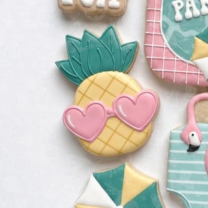 Puede incluir: Una colección de galletas de azúcar decoradas con temática veraniega. Una galleta es una piña amarilla con gafas de sol rosas en forma de corazón. Otras galletas incluyen un flamenco, una sombrilla y un diseño de playa.