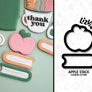 Peut inclure: Un ensemble d'emporte-pièces pour faire des biscuits en forme de pile de pommes. L'ensemble comprend un emporte-pièce en forme de pomme et un emporte-pièce en forme de pile de livres. Les emporte-pièces sont noirs et sont sur un fond blanc avec un motif d'étoiles, de cœurs et de notes de musique.