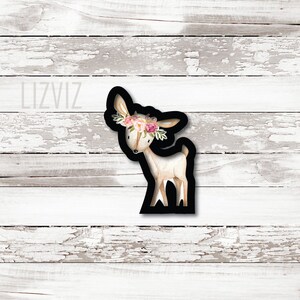 May include: A black and white cookie cutter in the shape of a deer with a floral crown. The deer is standing on all four legs and has a long neck. The floral crown is made of pink and white flowers.