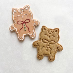 May include: Two cat-shaped cookies. One cookie is decorated with white icing outlines, a red icing bow, and three green icing dots. The other cookie is plain, showing the textured, light brown baked dough with the same cat shape and embossed details.
