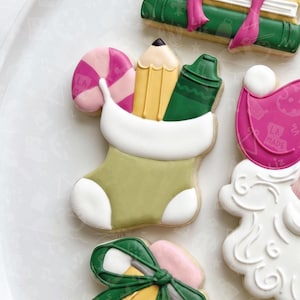 May include: A sugar biscuit shaped like a stocking with a white cuff, filled with a yellow pencil, a green crayon, and a pink and white striped sweet. The biscuit features smooth icing in olive green, white, yellow, pink, and dark green colours.