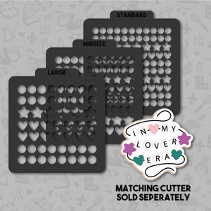 May include: Three black plastic stencils with different sizes labeled "Large", "Midsize", and "Standard". Each stencil has a grid of circles, hearts, and stars. The stencils are for crafting and decorating. The text "IN-MY LOVER ERA" is written in a decorative font with hearts and stars.
