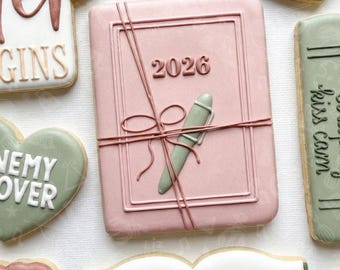 May include: A pink cookie in the shape of a planner, with the year 2026 embossed on the cover. A green pen and brown string are also on the cookie. Other cookies include the words "January", "Enemy Over", and "Kiss Calm".