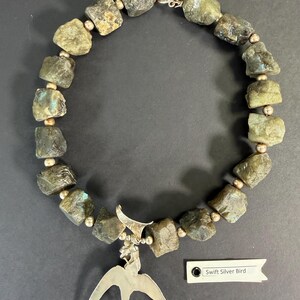 May include: A necklace featuring rough-cut, gray-green stones interspersed with small, gold-colored beads. A silver bird pendant hangs from the center. The tag reads "Swift Silver Bird".