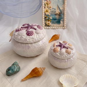 Op de afbeelding: Twee ronde witte keramieken sieradendoosjes met een textuur, elk bekroond met een paarse zeester en diverse schelpen. Een rechthoekige fotolijst toont een zeilboot op het water met bloemen. Losse schelpen liggen verspreid op de ondergrond.