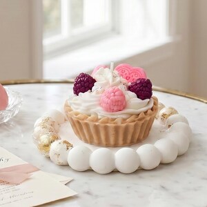 Könnte beinhalten: Eine dessertförmige Kerze mit einem beigefarbenen Tortenboden, verziert mit weißen Wachswirbeln, beerenförmigen Wachsstücken in Pink und Dunkellila sowie einem herzförmigen rosa Wachsakzent auf einem weißen, runden Tablett mit perlenartigem Rand.