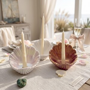 Peut inclure: Trois bougeoirs en forme de coquillage aux finitions blanche, irisée et cuivre métallique, chacun contenant une longue bougie effilée de couleur crème. L'ensemble est disposé sur une surface en tissu clair avec un petit coquillage vert.