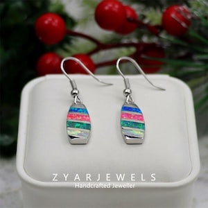 May include: A pair of silver earrings with a colorful, striped design. Each earring features a rectangular shape with horizontal bands of blue, pink, green, and white. The earrings are displayed on a white surface, with the text "ZYAR JEWELS Handcrafted Jeweller" visible.