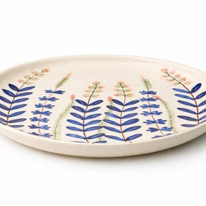 May include: A cream-colored ceramic plate with a hand-painted botanical design. The design features blue, leaf-like shapes with pink and green accents. The plate is round and has a slightly raised edge, perfect for serving food.