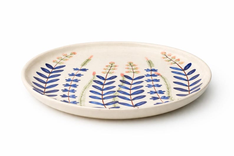 May include: A cream-colored ceramic plate with a hand-painted botanical design. The design features blue, leaf-like shapes with pink and green accents. The plate is round and has a slightly raised edge, perfect for serving food.