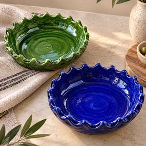Op de afbeelding: Twee ronde keramische schalen met geschulpte, golvende randen. De ene schaal is diep, glanzend bosgroen en de andere is levendig, glanzend kobaltblauw. Beide stukken hebben een gladde, reflecterende afwerking en een ronde, verzonken basis.