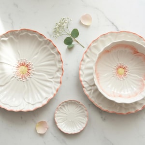 Puede incluir: Juego de vajilla de cerámica blanca con diseño de flor de amapola. El juego incluye platos grandes, cuencos y un plato pequeño, todos con borde y centro rosa. Una ramita de flores blancas y pétalos están dispuestos cerca.