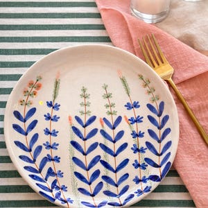 May include: A white ceramic plate with a hand-painted floral design featuring blue leaves and small blue and pink flowers. The plate is set on a green and white striped tablecloth, with a pink napkin and gold fork.