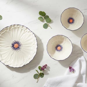 May include: A white ceramic dinnerware set with a floral design. The set includes a large plate, bowls, and a teacup, all with a blue rim and a central orange and blue flower. The set is arranged on a white surface.