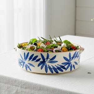 May include: A ceramic serving bowl filled with a fresh salad. The bowl has a cream base with a blue floral design. The salad includes tomatoes, olives, red onion, and feta cheese. Rosemary sprigs and basil leaves garnish the salad.