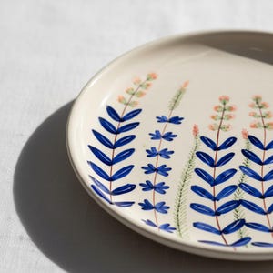 May include: A white ceramic plate with a hand-painted floral design. The design features blue leaf-like shapes and small pink and green floral accents. The plate has a slightly raised edge and is set against a white background.