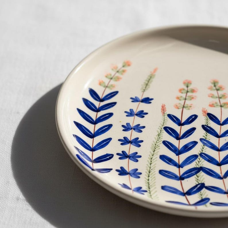 May include: A white ceramic plate with a hand-painted floral design. The design features blue leaf-like shapes and small pink and green floral accents. The plate has a slightly raised edge and is set against a white background.