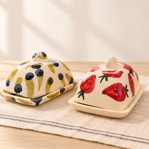 May include: Two ceramic butter dishes with lids. One dish features a hand-painted pattern of blue berries and green leaves on a cream background. The other dish displays a hand-painted pattern of red strawberries with green stems on a cream base.