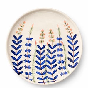 May include: A round, off-white ceramic plate with a hand-painted floral design. The design features blue leaves and stems, with pink and green flower clusters. The plate has a slightly raised edge and a smooth surface.