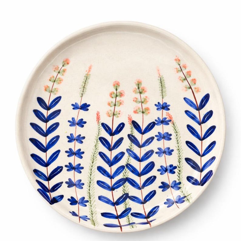May include: A round, off-white ceramic plate with a hand-painted floral design. The design features blue leaves and stems, with pink and green flower clusters. The plate has a slightly raised edge and a smooth surface.