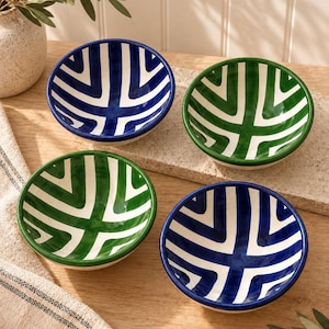 May include: A set of four round ceramic bowls featuring a hand-painted geometric pattern of white lines against solid blue or green backgrounds. The bowls have a smooth, glossy finish and are arranged in a square formation on a wooden surface.