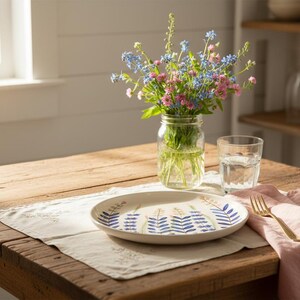 May include: A rustic wooden table is set with a floral arrangement in a mason jar, a plate with a blue and yellow design, a glass of water, and a pink napkin with a gold fork. The scene is bathed in natural light.