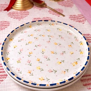 May include: Two white ceramic plates with a floral pattern of small yellow and pink flowers. The plates have a blue border with a dotted design. The plates are stacked on a pink and white heart-patterned tablecloth.