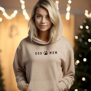 May include: A sand-colored hoodie with the text "DOG MOM" and a paw print graphic. The hoodie has a hood and a front pocket. The model is wearing the hoodie.