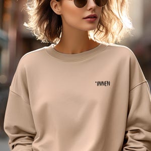 Könnte beinhalten: Ein beigefarbenes Sweatshirt mit dem Wort "*INNEN" in Schwarz auf der Brust gedruckt. Das Sweatshirt hat einen Rundhalsausschnitt und lange Ärmel. Die Person auf dem Bild trägt eine Sonnenbrille und blaue Jeans.