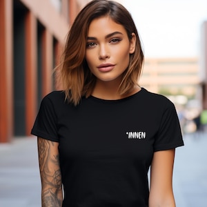 Könnte beinhalten: Schwarzes T-Shirt mit Rundhalsausschnitt und dem Wort "*INNEN" in Weiß auf der Brust. Das Shirt besteht aus einem weichen Material und hat kurze Ärmel. Das Model trägt eine blaue Jeans.