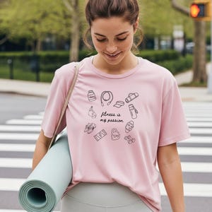 Könnte beinhalten: Hellrosa T-Shirt mit dem Schriftzug "fitness is my passion" und Fitness-Symbolen. Die Person hält eine zusammengerollte Yogamatte. Im Hintergrund ein Zebrastreifen und Bäume.