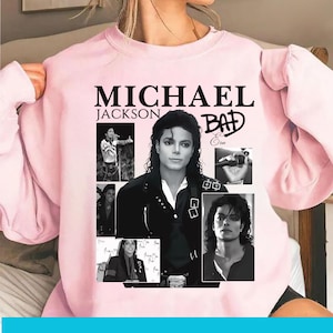May include: Pale pink sweatshirt with a collage of images of Michael Jackson. The design includes the text "MICHAEL JACKSON BAD" in black. The collage includes black and white photos of the singer.