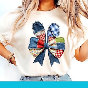 May include: Cream-colored t-shirt featuring a patchwork bunny ear design. The design incorporates denim, red gingham, floral, and blue gingham fabrics, creating a textured and colorful Easter-themed graphic.