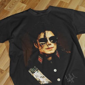 Pode incluir: Camiseta preta com um retrato de Michael Jackson usando &oacute;culos de sol. A imagem &eacute; um close-up do rosto do cantor, com sua assinatura em branco no canto inferior direito. A camiseta est&aacute; estendida em uma superf&iacute;cie de madeira.