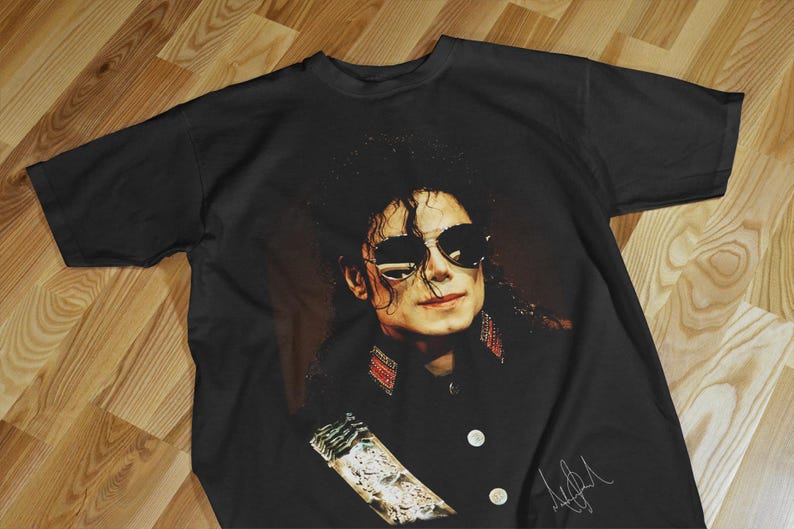 Pode incluir: Camiseta preta com um retrato de Michael Jackson usando &oacute;culos de sol. A imagem &eacute; um close-up do rosto do cantor, com sua assinatura em branco no canto inferior direito. A camiseta est&aacute; estendida em uma superf&iacute;cie de madeira.