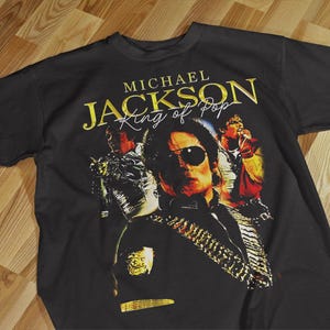 Pode incluir: Camiseta preta com um gr&aacute;fico dourado de Michael Jackson usando &oacute;culos de sol e o texto "MICHAEL JACKSON King of Pop". O design inclui imagens de Jackson em v&aacute;rias poses.