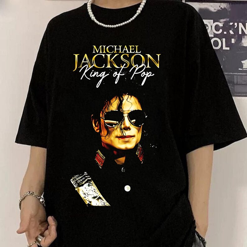 May include: Black t-shirt with a gold print of Michael Jackson's portrait and the text "MICHAEL JACKSON King of Pop". The short-sleeved shirt has a crew neck. The image shows a close-up of the design.