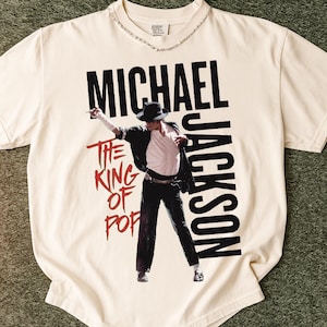 May include: Cream-coloured t-shirt featuring a graphic of Michael Jackson in a black suit and hat. The text "MICHAEL JACKSON" is printed in black, with "THE KING OF POP" in red. The shirt has a crew neck with a silver chain.