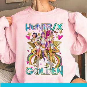 Peut inclure: Sweat-shirt rose clair avec l'inscription "HUNTXRX GOLDEN" en turquoise et or. Le graphisme comprend trois personnages stylisés, une étoile dorée, des cœurs et des arcs-en-ciel. Les personnages portent des tenues blanches et dorées.