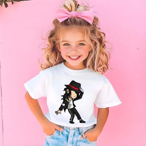 May include: A white t-shirt with a cartoon illustration of a person in a black hat and suit, with a red stripe on the hat. The person is in a dance pose. The shirt is worn by a young person with a pink bow in their hair.