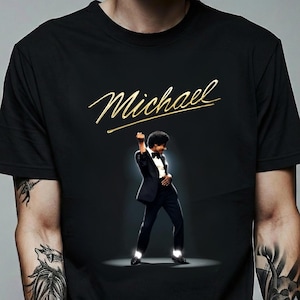 May include: Black t-shirt with the gold script name "Michael" above an image of a man in a black tuxedo, striking a pose. The image is set against a dark background, with a spotlight effect.