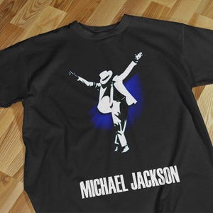 Pode incluir: Camiseta preta com uma silhueta branca de Michael Jackson em uma pose de dan&ccedil;a, com os bra&ccedil;os levantados. O texto "MICHAEL JACKSON" &eacute; impresso em branco abaixo da imagem. Um efeito de respingo azul est&aacute; atr&aacute;s da figura.