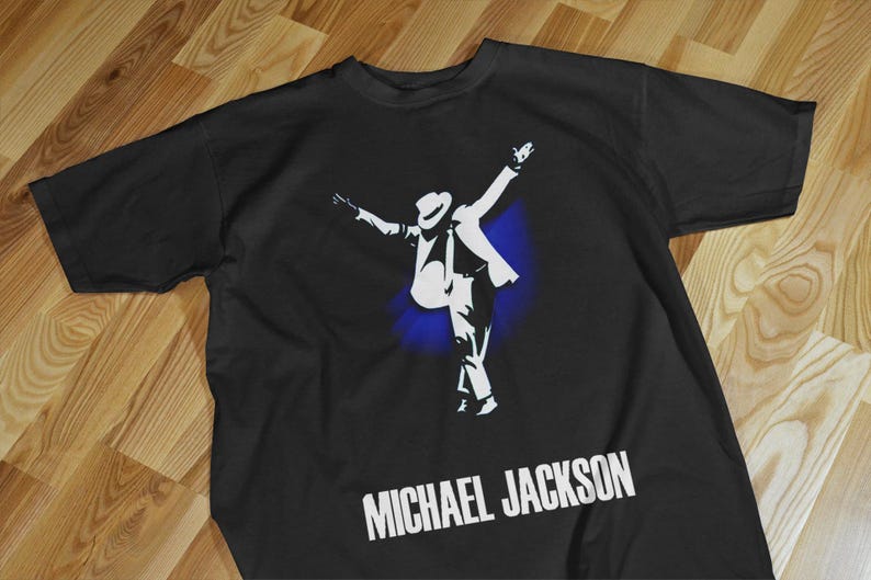 Pode incluir: Camiseta preta com uma silhueta branca de Michael Jackson em uma pose de dan&ccedil;a, com os bra&ccedil;os levantados. O texto "MICHAEL JACKSON" &eacute; impresso em branco abaixo da imagem. Um efeito de respingo azul est&aacute; atr&aacute;s da figura.