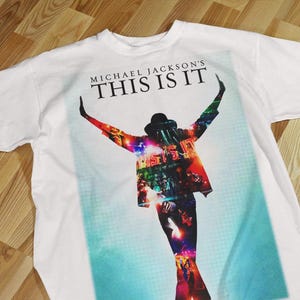 Pode incluir: Camiseta branca com um gr&aacute;fico colorido de Michael Jackson com os bra&ccedil;os levantados e o texto "MICHAEL JACKSON'S THIS IS IT" no topo. A imagem tem um esquema de cores azul e vermelho.