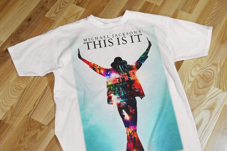 Pode incluir: Camiseta branca com um gr&aacute;fico colorido de Michael Jackson com os bra&ccedil;os levantados e o texto "MICHAEL JACKSON'S THIS IS IT" no topo. A imagem tem um esquema de cores azul e vermelho.