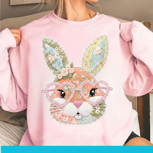 Puede incluir: Sudadera rosa claro con un diseño de conejo patchwork. El conejo tiene gafas en forma de corazón, orejas y nariz con estampado floral. El diseño utiliza una variedad de colores y patrones.