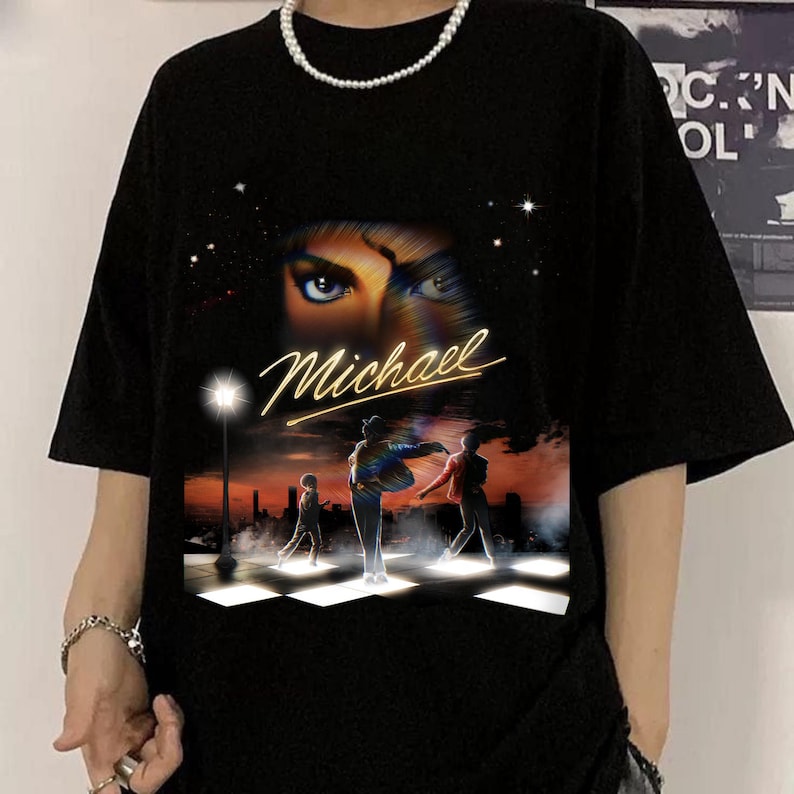 May include: Black t-shirt with a graphic of Michael Jackson's eyes and the name "Michael" in gold script. The graphic includes a scene of figures crossing a street at sunset. The shirt has short sleeves and a crew neck.